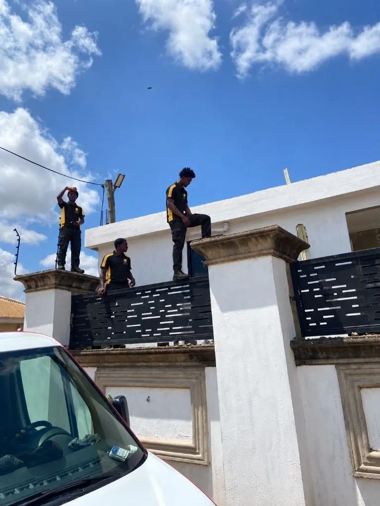 Three JakShield technicians in uniform working on top of a compound wall and gate pillars, installing an electric fence.