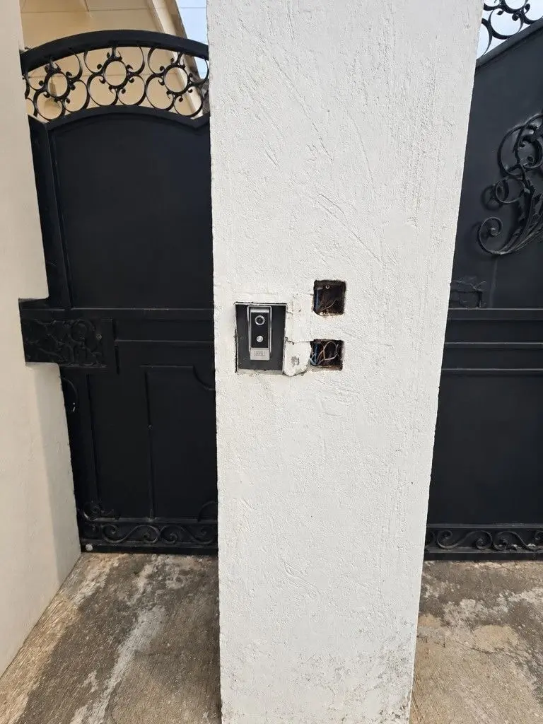 Video intercom door station mounted on a white pillar beside a black gate.