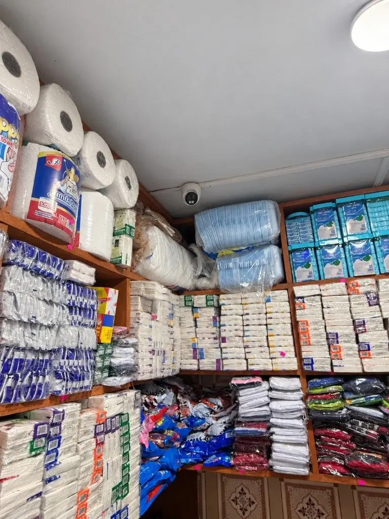 Turret CCTV camera on the ceiling of a provision shop, above shelves stocked with tissue, napkins and household goods.