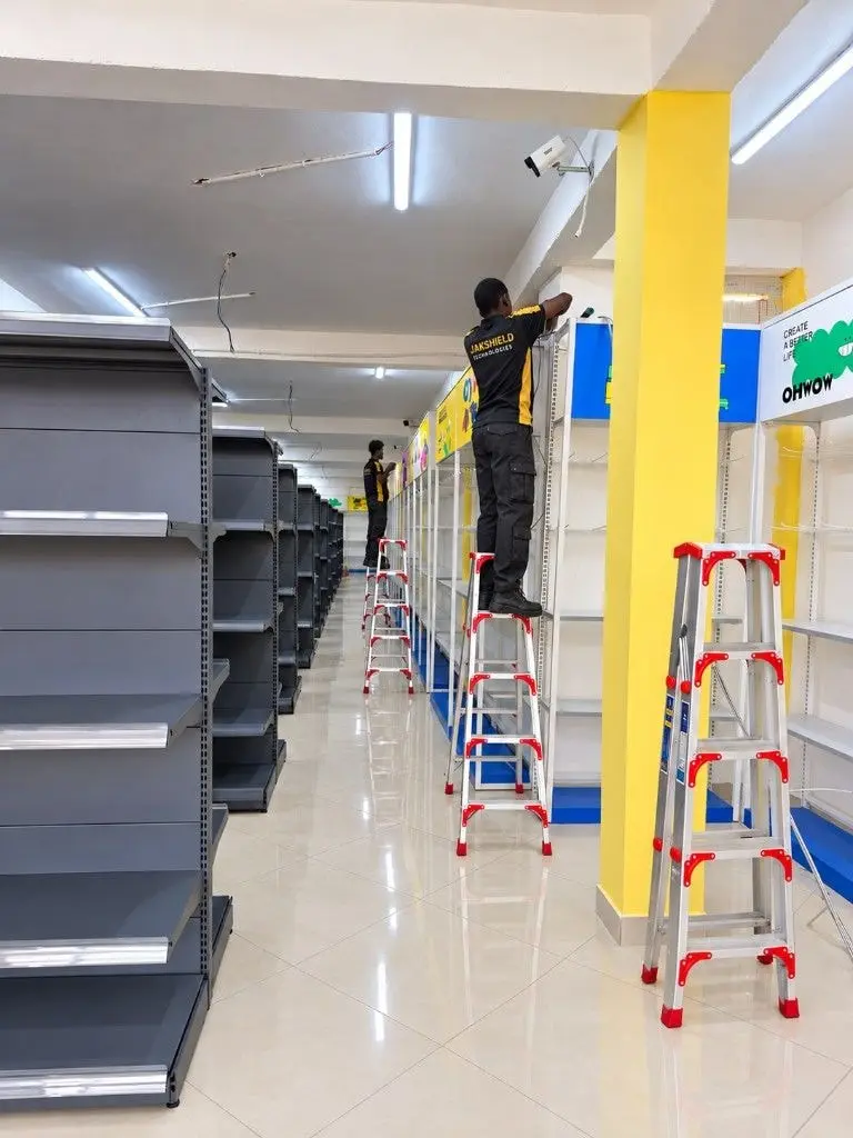 JakShield technicians in uniform on ladders fitting CCTV cameras along the ceiling of a large new retail store with empty shelves.