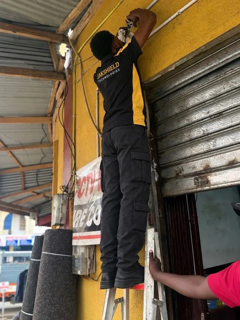 A JakShield technician on a ladder fixing a CCTV camera above the roller shutter of a shop.